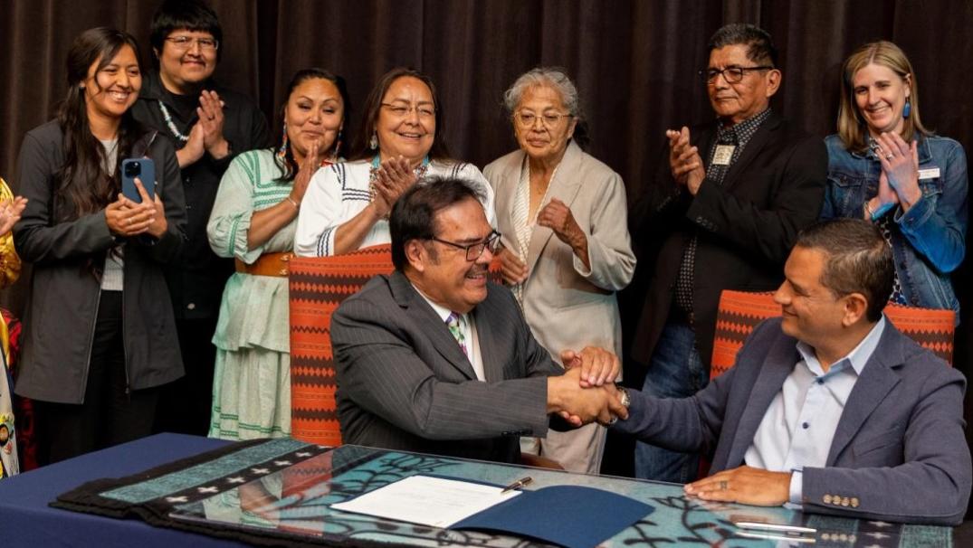 Photo of NAU President Cruz Rivera and SCAC President Martin Ahumada shaking hands at a table surrounded by clapping people. 