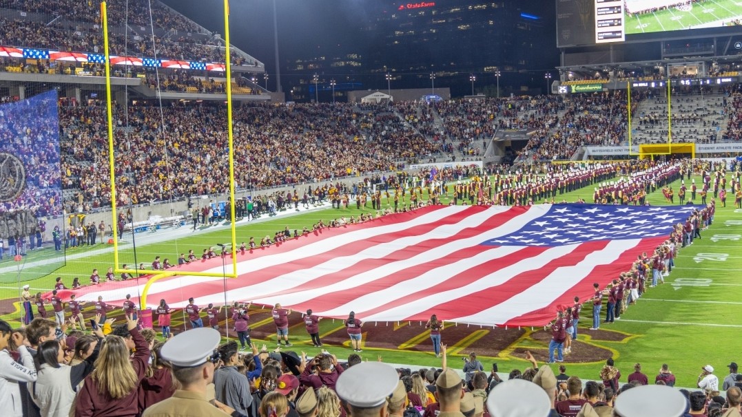 Image of a huge American flag carried by people on ASU's football field with military members and civilians in the audience. 