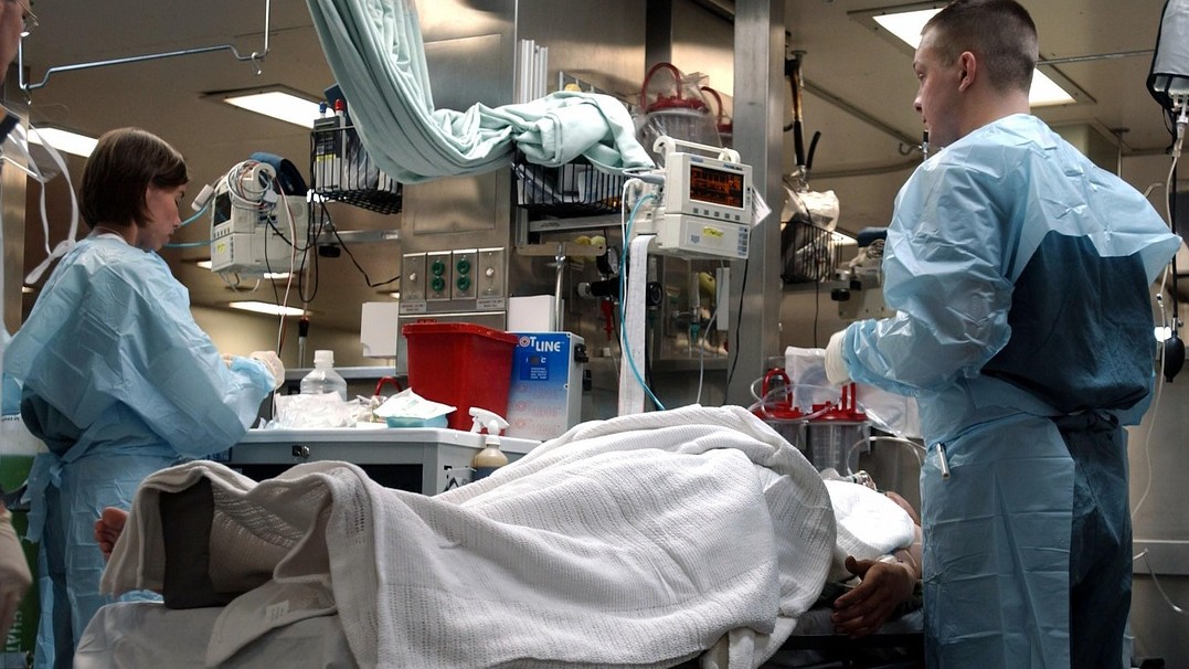 An hospital surgical suite is pictured with a man and woman in scrubs working on a patient who is covered in a blanket. 