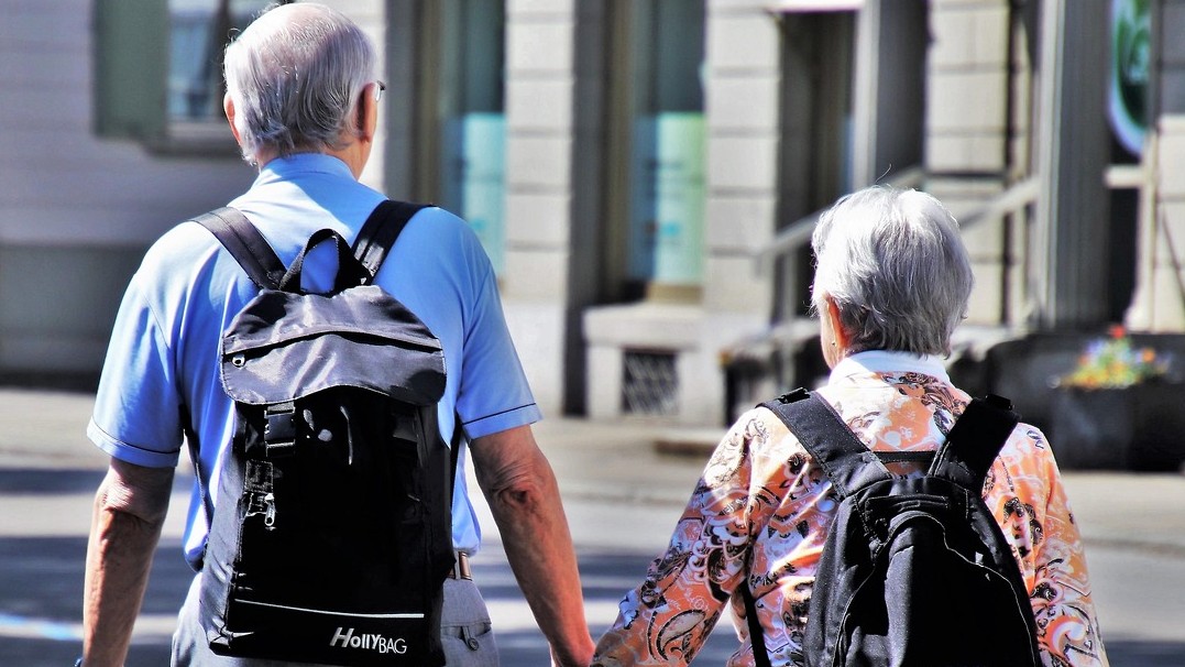 Photo of an elderly man and woman shot from behind who are holding hands. 