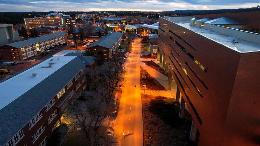 Aerial image of the NAU campus at dusk with lights showing in buildings and lights along the pedway.