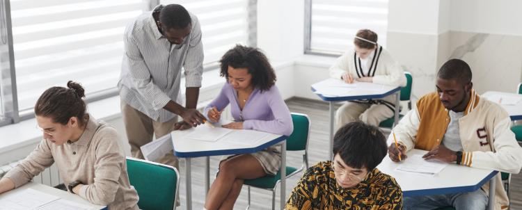 Photo of a teacher in a classroom helping a female student while other students work 