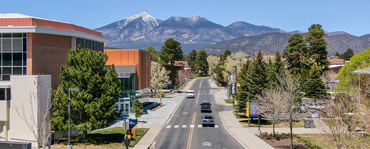 Image of NAU campus with mountains in the background and a road in the middle of the photos as well as buildings to the left 